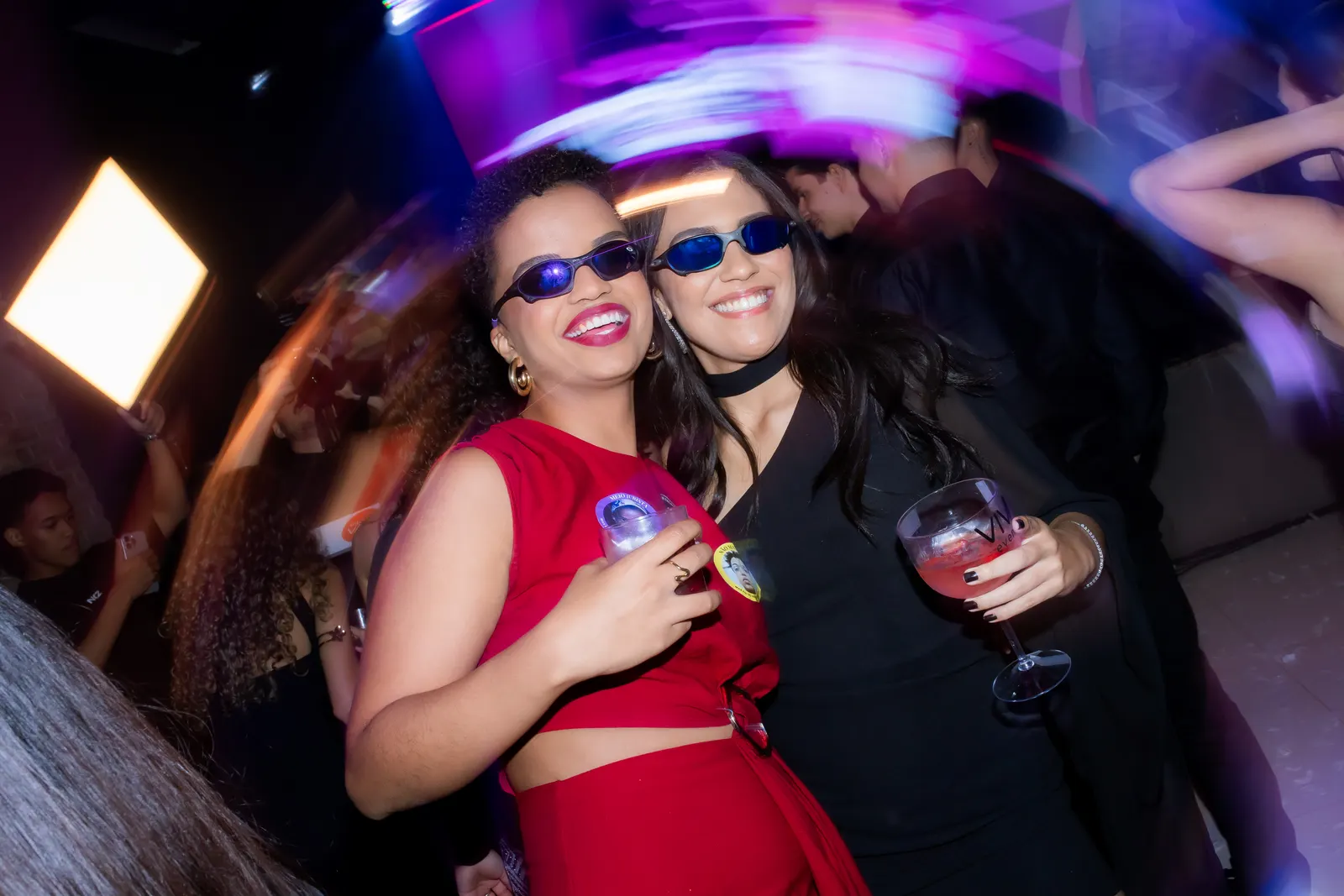 Duas formandas de óculos escuros dançando na pista de uma festa de pré-evento, com luzes coloridas em movimento.