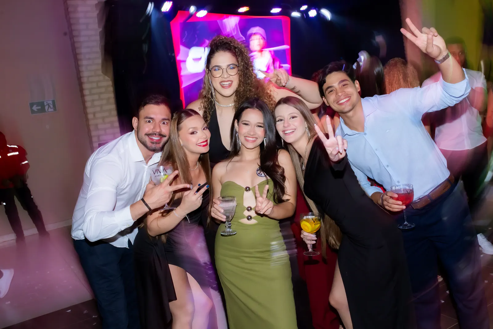 Grupo de formandos sorrindo e fazendo sinais com as mãos em frente ao palco da festa.