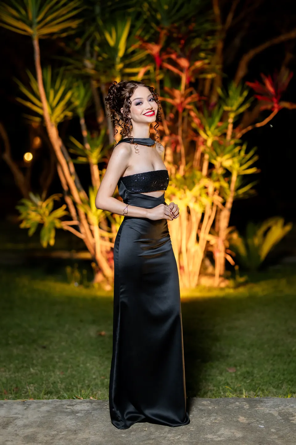 Formanda de vestido longo preto posando à noite num jardim iluminado.