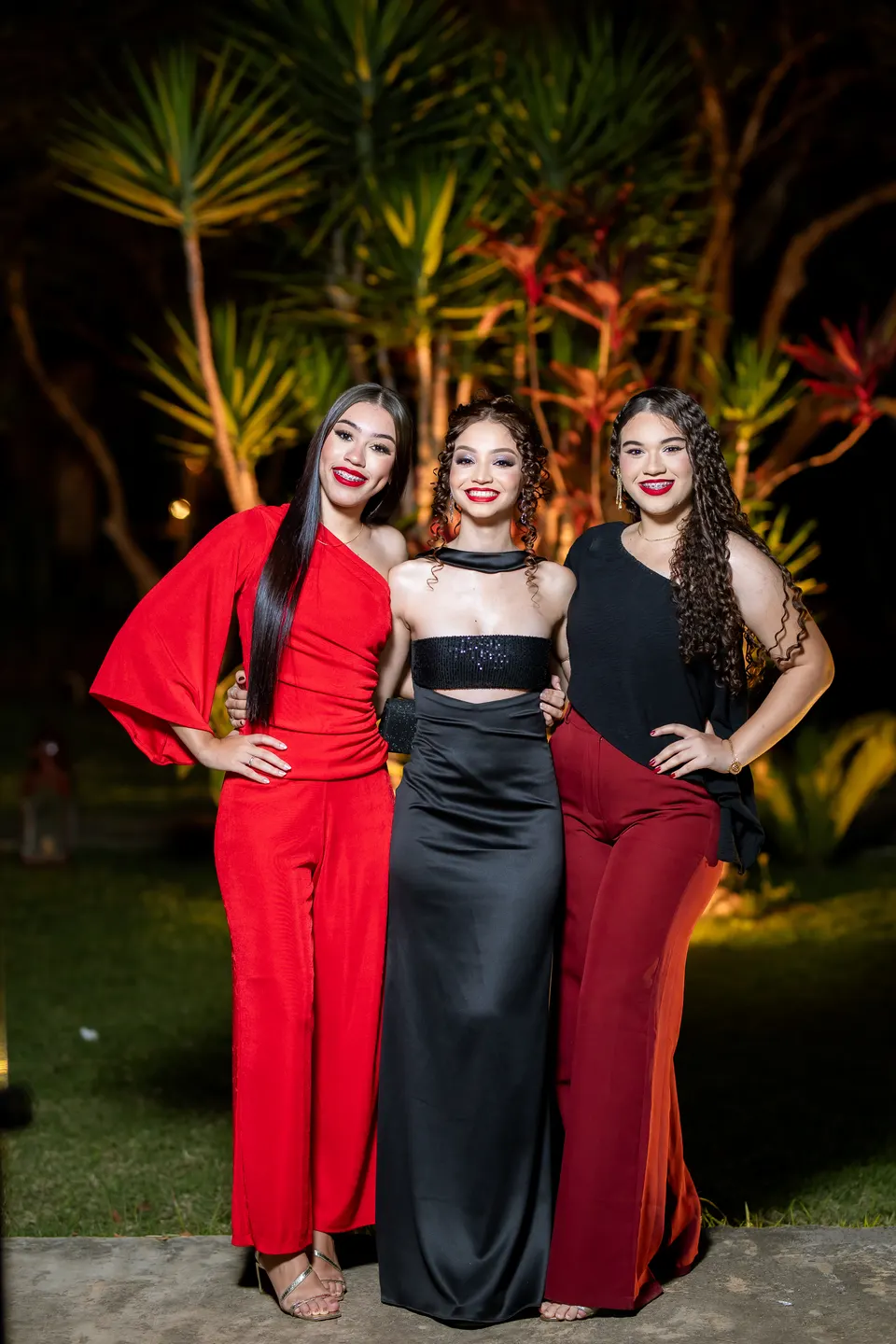 Três formandas de vestidos longos, dois vermelhos e um preto, posando juntas no jardim à noite.