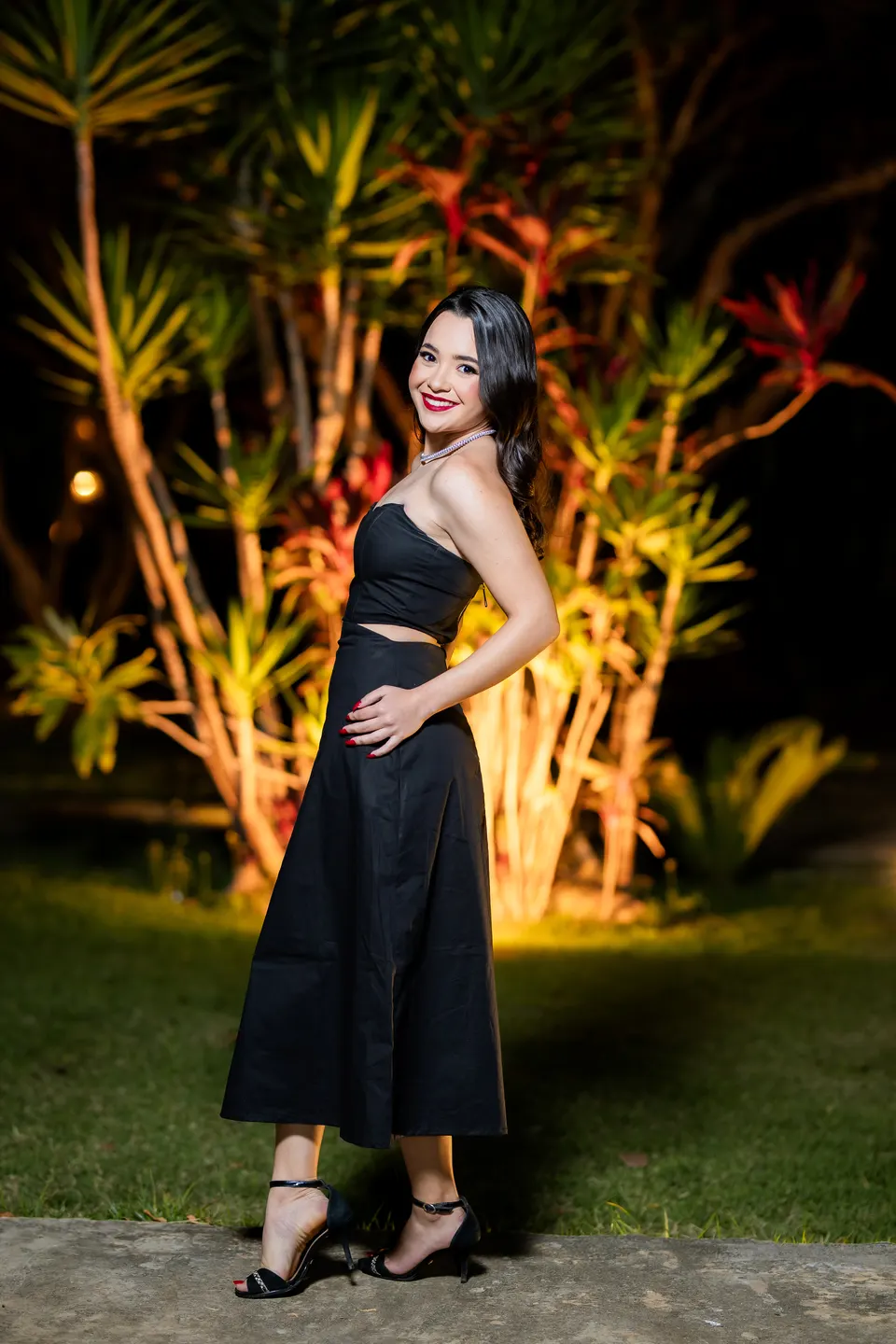 Formanda de vestido preto sorrindo em retrato noturno no jardim iluminado.