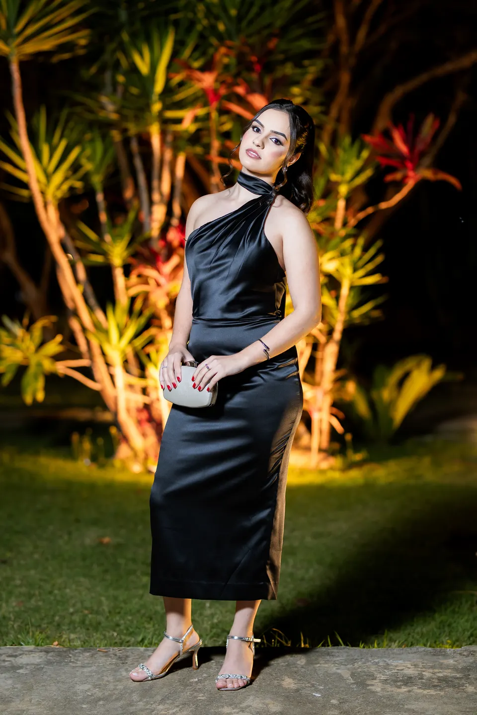 Formanda de vestido preto acetinado posando com a bolsa de mão no jardim iluminado.