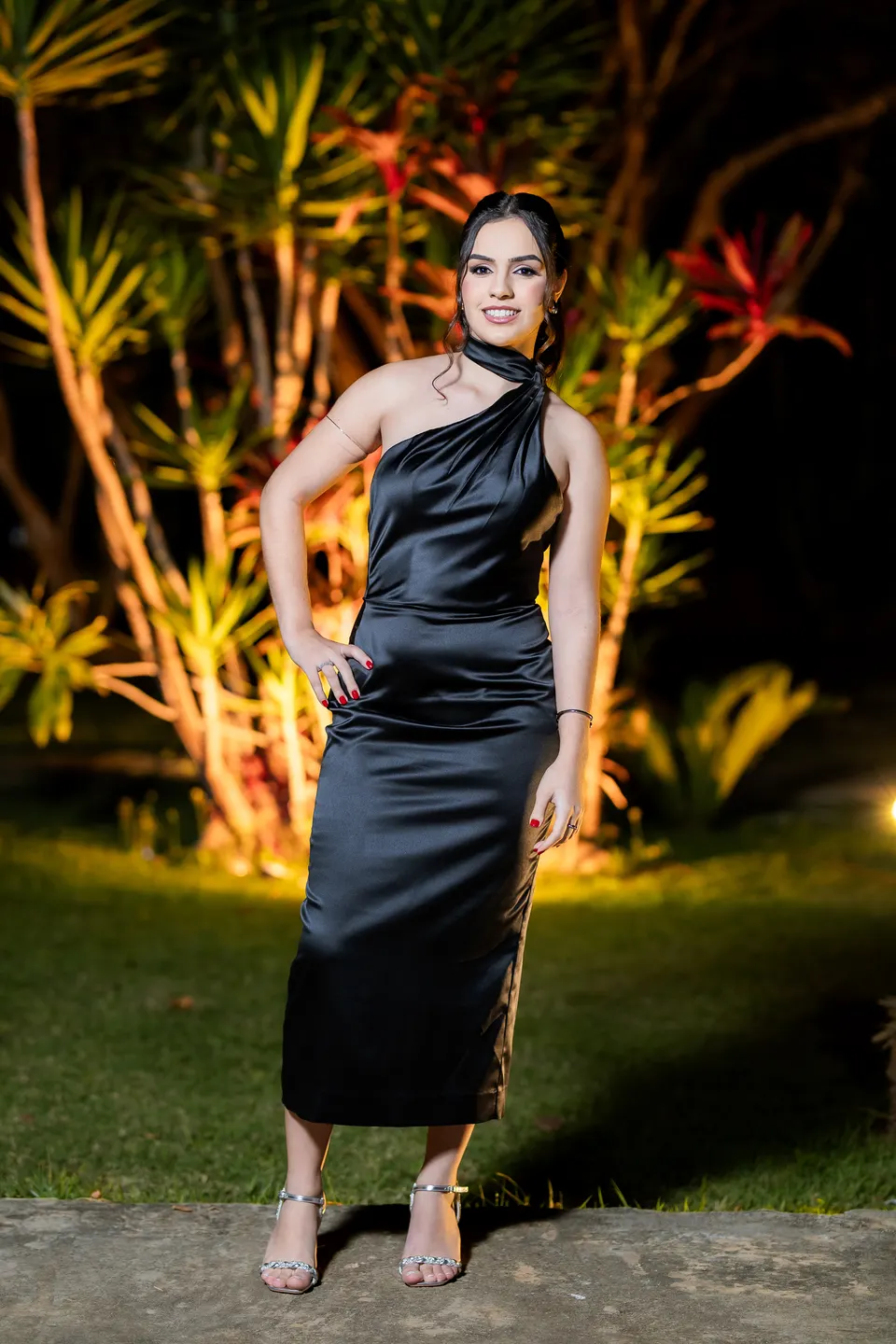 Formanda de vestido preto de um ombro só, com a mão na cintura, no jardim à noite.