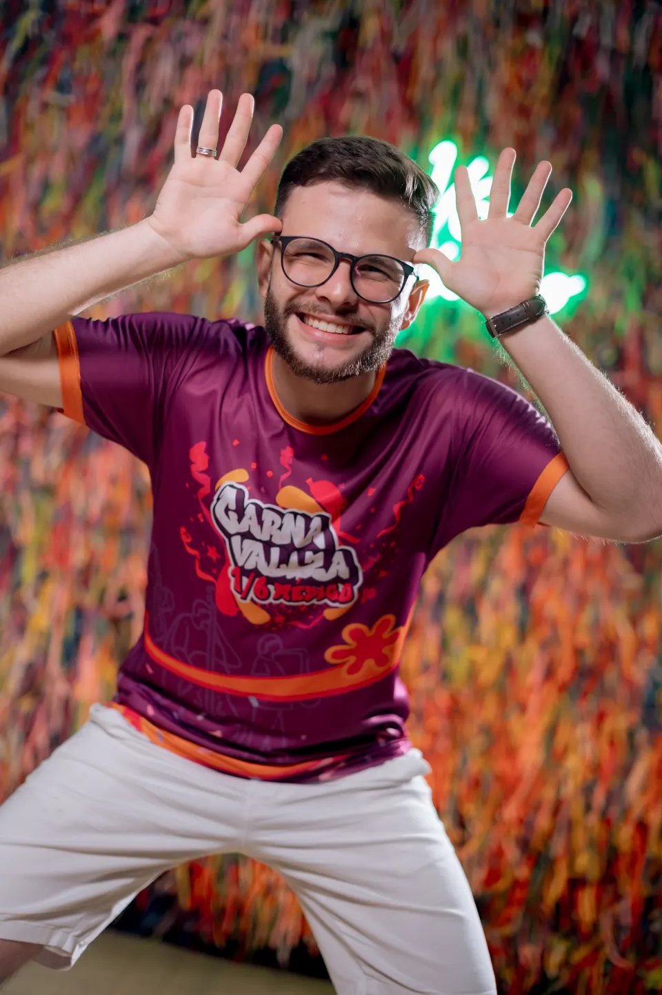 Formando de óculos fazendo careta com as mãos abertas em frente ao painel de fitas coloridas.