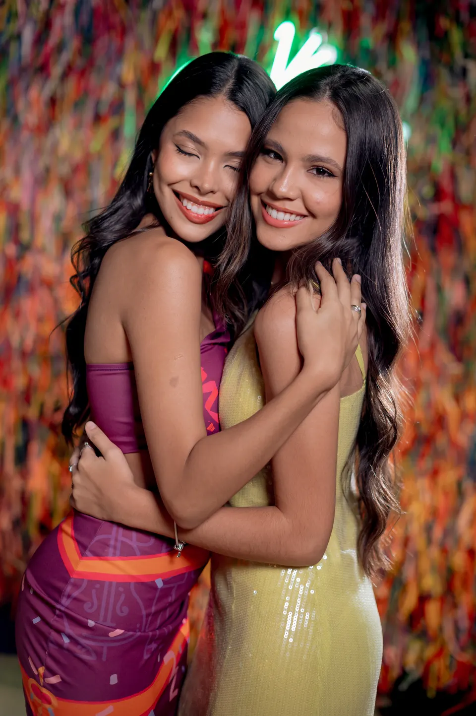 Duas formandas abraçadas e sorrindo em frente ao painel de fitas coloridas.