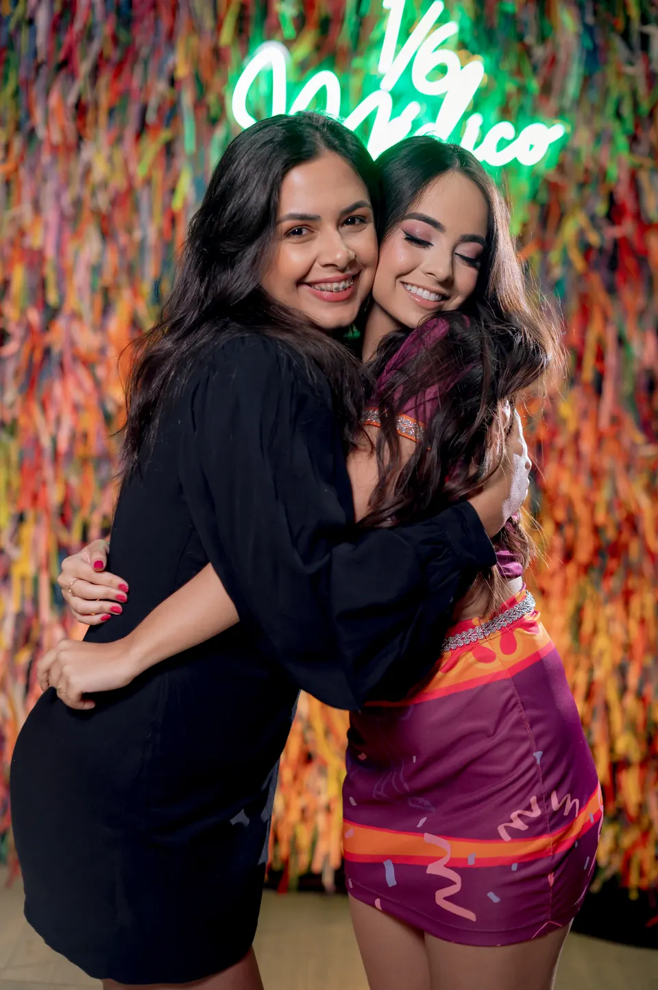 Duas amigas abraçadas sorrindo em frente ao painel de fitas com letreiro em neon.