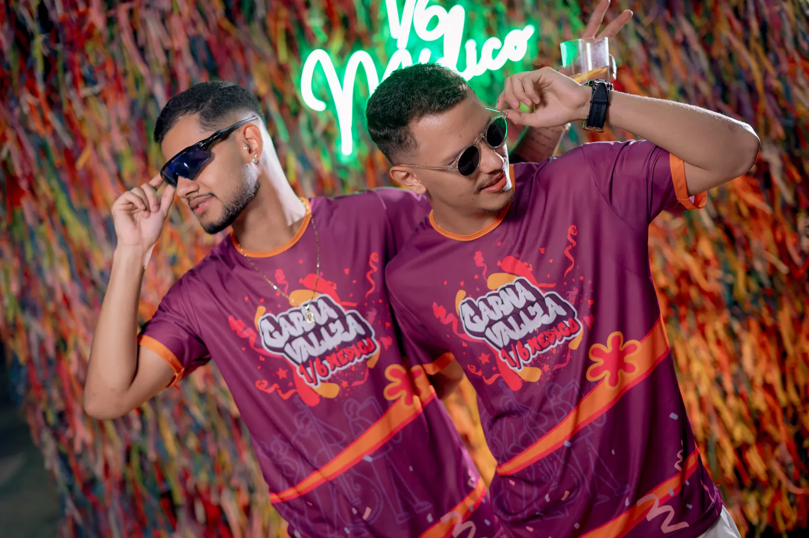 Dois formandos de óculos escuros com a camisa da festa Carnavalizza fazendo pose em frente ao painel neon.