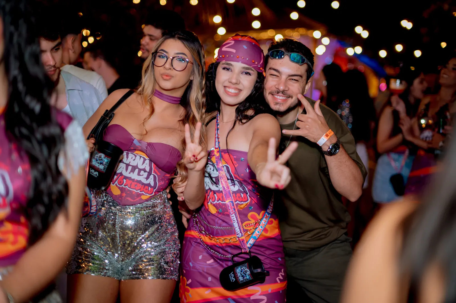 Três amigos fazendo sinal de paz na festa, sob um varal de luzes.
