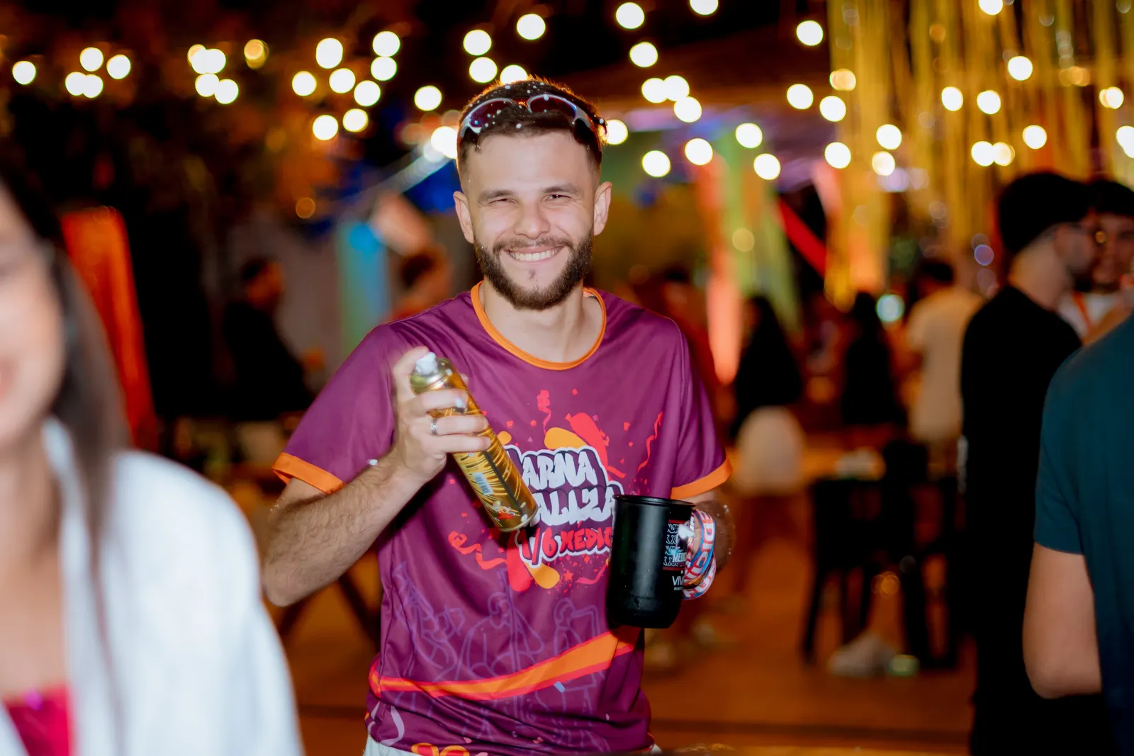 Formando sorrindo com a camisa da festa e uma garrafa na mão, sob um varal de luzes.