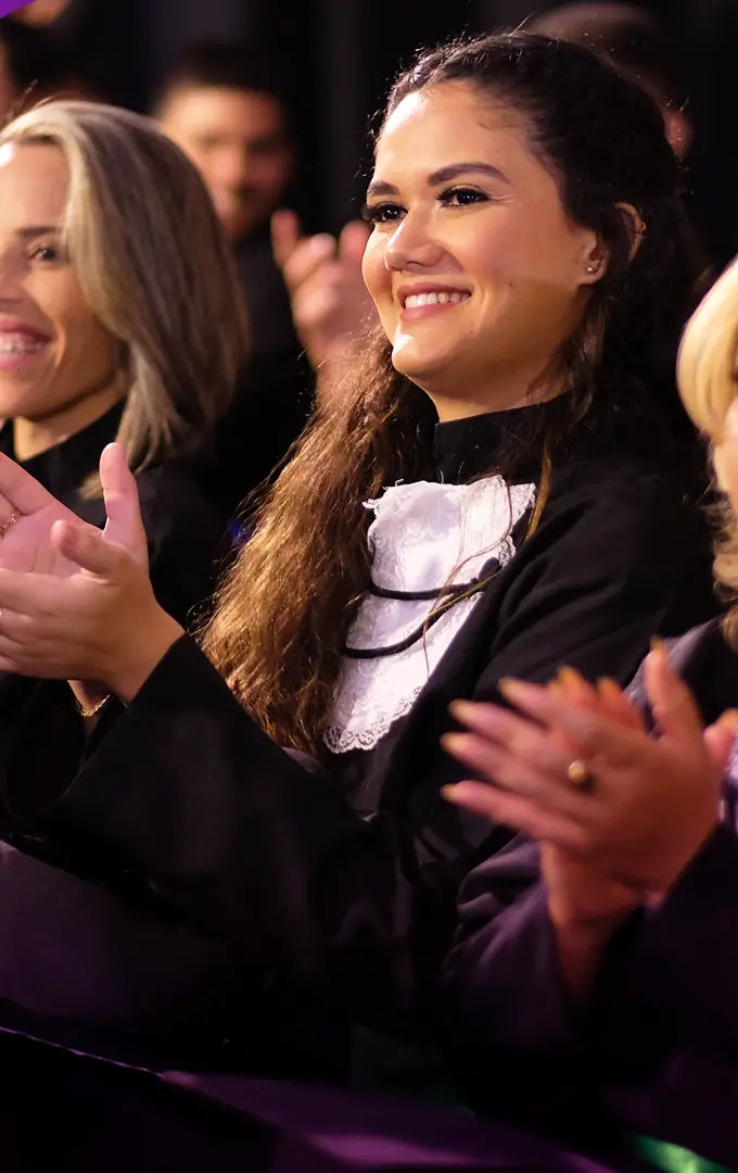 Formanda de beca aplaude sentada na plateia durante a cerimônia, ao lado de outras convidadas.
