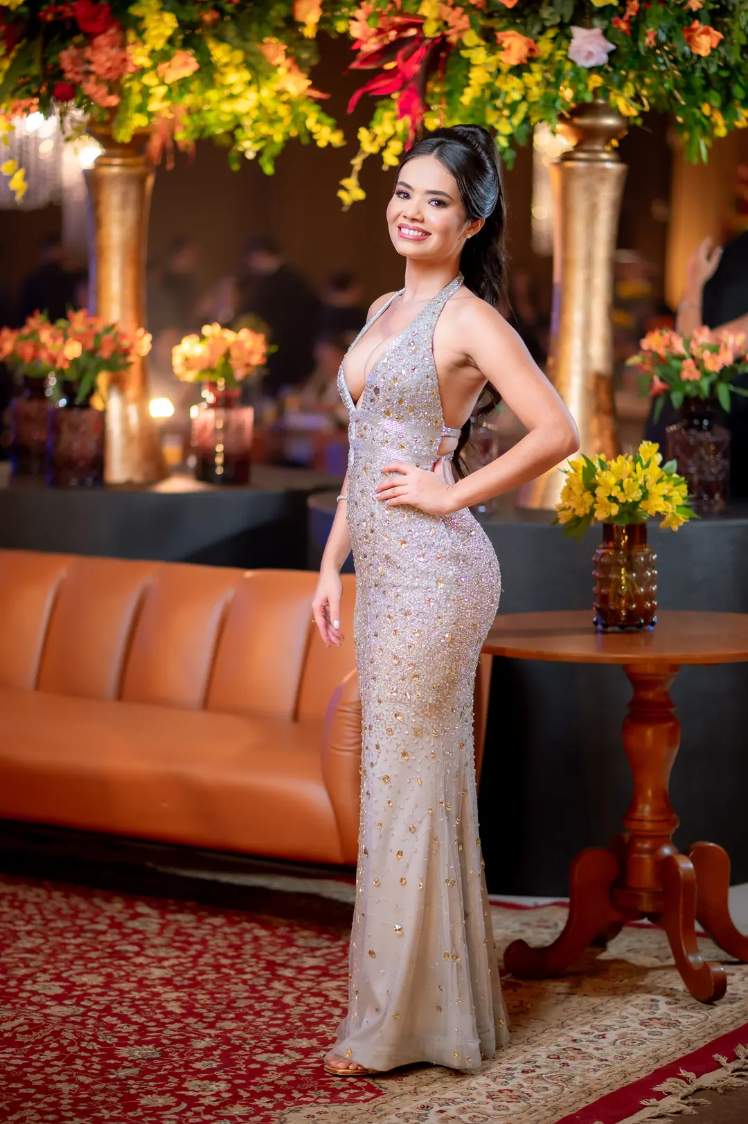 Formanda em vestido longo prateado no salão do baile de gala, sob a decoração floral suspensa.