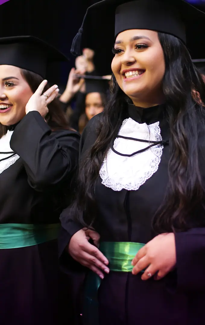Duas formandas de beca com faixa verde ajustam o capelo e sorriem antes da entrada da cerimônia.