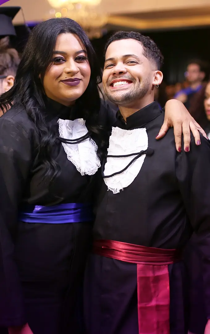 Dois formandos de becas com faixas de cores diferentes, de cursos distintos, abraçados e sorrindo durante a colação unificada.