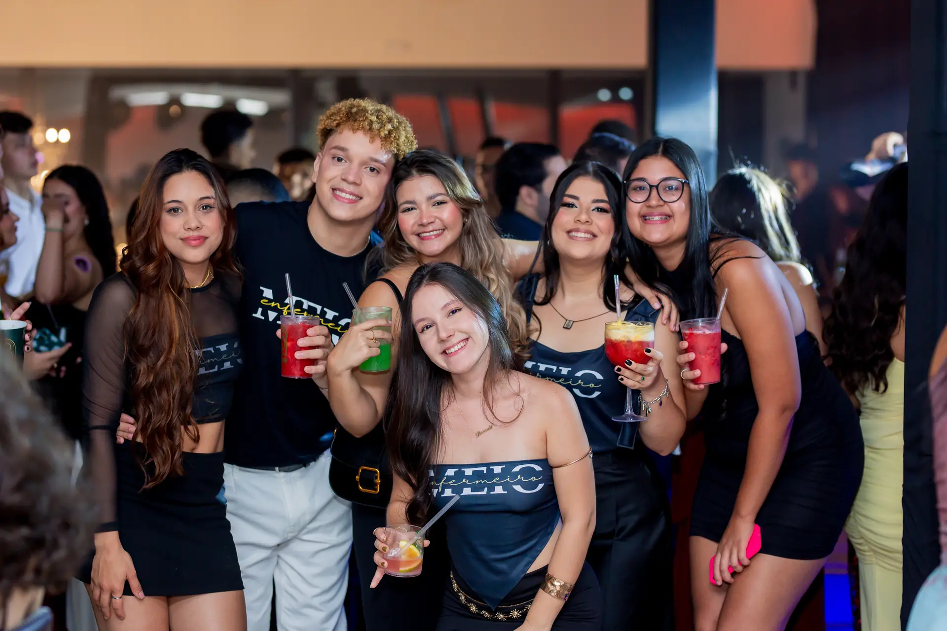 Quatro estudantes de camiseta da turma brindam com drinks coloridos na festa de meio de curso.